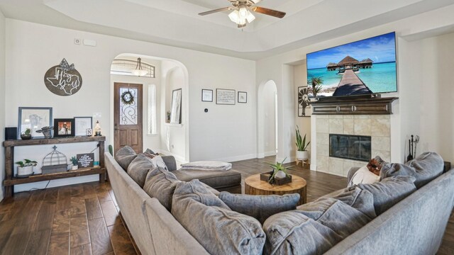 living area with a tiled fireplace, arched walkways, dark wood-type flooring, and a tray ceiling