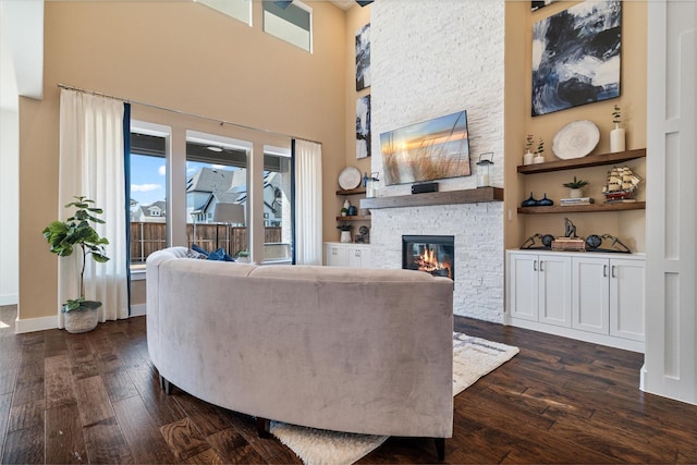 living area with a stone fireplace, dark wood-style flooring, a towering ceiling, and baseboards