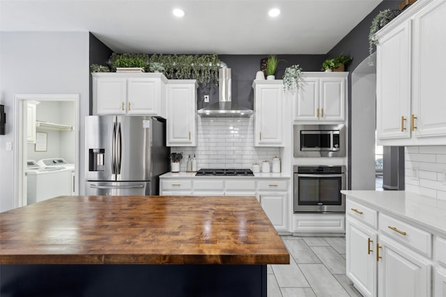 kitchen featuring wall chimney exhaust hood, butcher block countertops, stainless steel appliances, washing machine and dryer, and white cabinetry