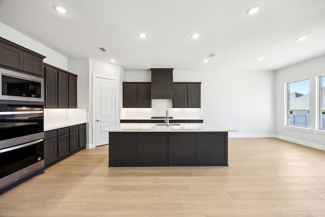 kitchen with black microwave, light wood-style flooring, a sink, light countertops, and an island with sink