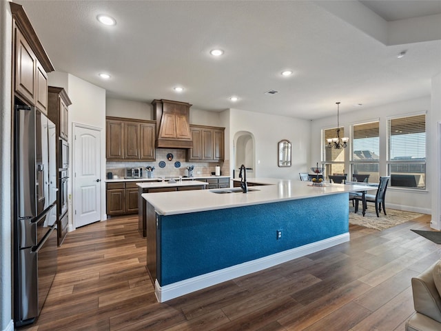 kitchen with dark wood-style flooring, a sink, stainless steel fridge with ice dispenser, a center island with sink, and custom range hood