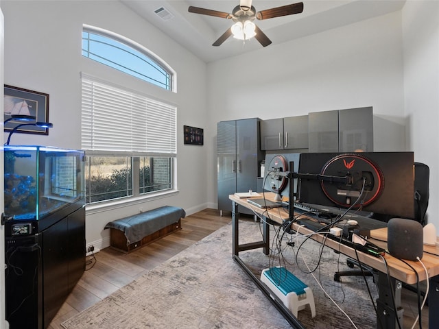 home office with light wood finished floors, visible vents, a high ceiling, a ceiling fan, and baseboards