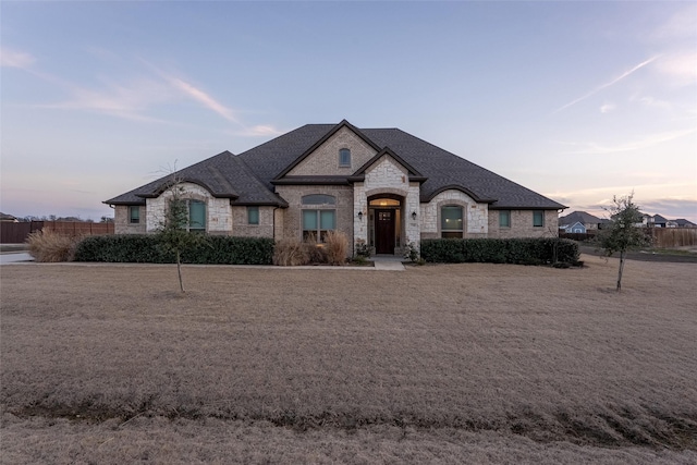 french country home with stone siding, a shingled roof, fence, and brick siding