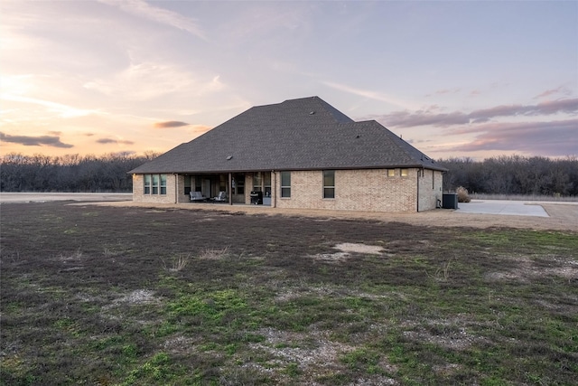 back of house at dusk featuring a patio, central AC unit, roof with shingles, and brick siding