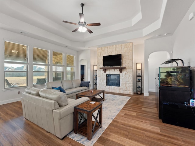 living room with a tray ceiling, visible vents, a stone fireplace, wood finished floors, and baseboards