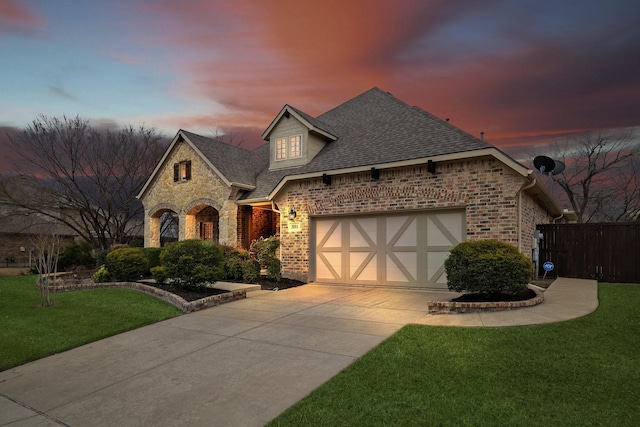 french country inspired facade featuring roof with shingles, a yard, brick siding, an attached garage, and driveway