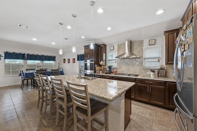 kitchen with visible vents, wall chimney range hood, appliances with stainless steel finishes, an island with sink, and decorative light fixtures