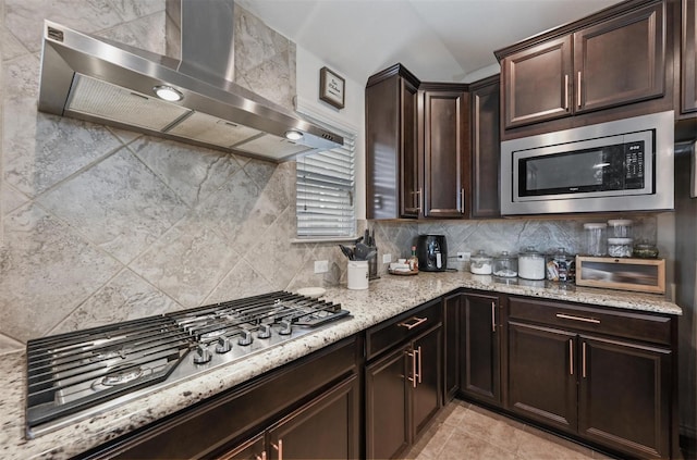 kitchen with wall chimney exhaust hood, light stone counters, stainless steel appliances, dark brown cabinets, and backsplash