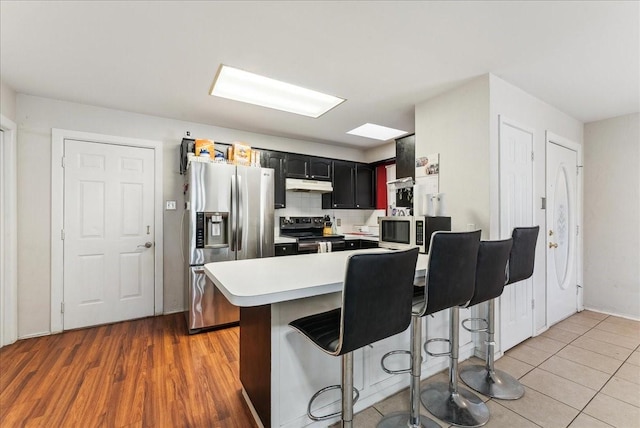 kitchen with a breakfast bar area, under cabinet range hood, light countertops, appliances with stainless steel finishes, and backsplash