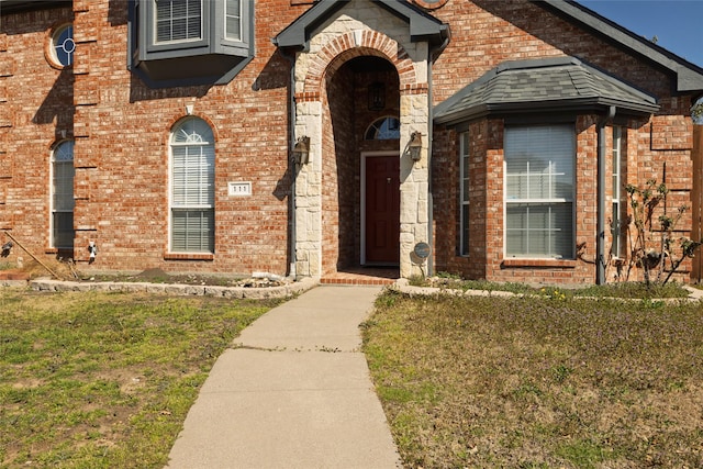 entrance to property featuring a shingled roof, brick siding, and a yard