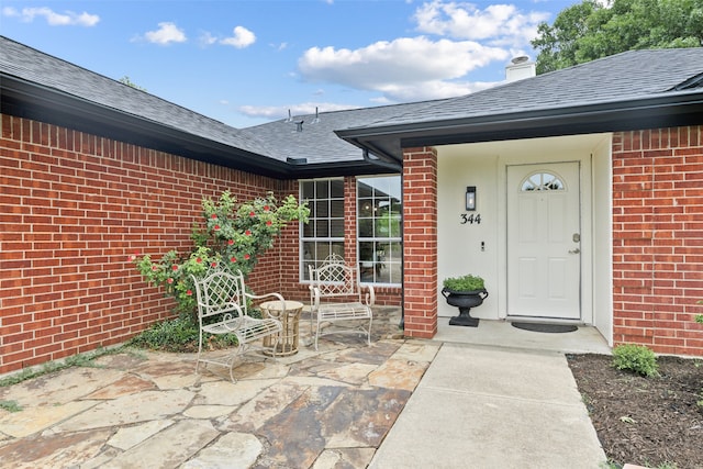 doorway to property featuring a shingled roof, a patio area, and brick siding