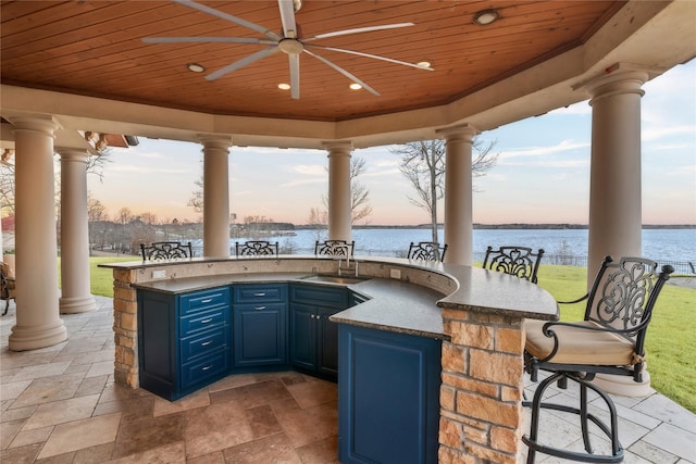 patio terrace at dusk featuring ceiling fan, outdoor wet bar, a water view, a sink, and area for grilling