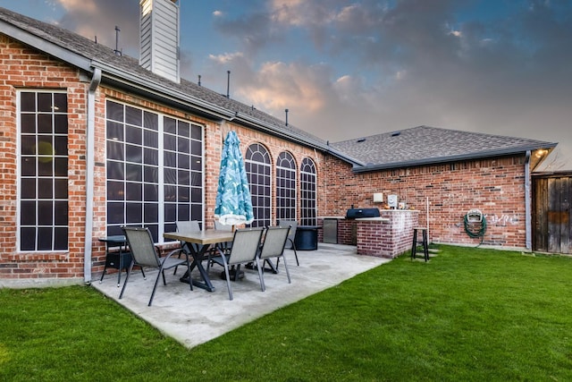 back of property at dusk with a lawn, a patio, roof with shingles, brick siding, and a chimney