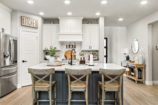 kitchen with stainless steel fridge, a kitchen island with sink, white cabinets, and light countertops