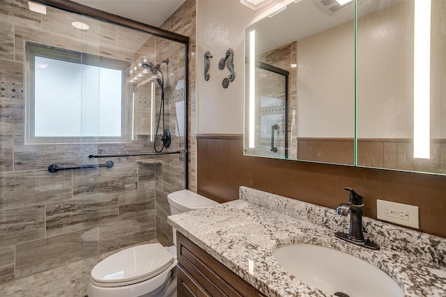 bathroom featuring a stall shower, toilet, a wainscoted wall, vanity, and tile walls