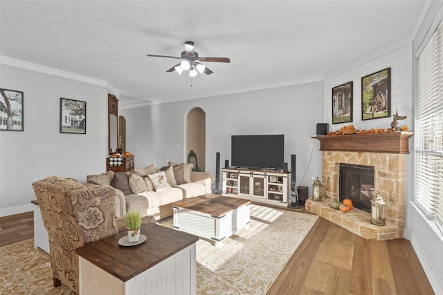 living area featuring arched walkways, ceiling fan, ornamental molding, wood finished floors, and a stone fireplace