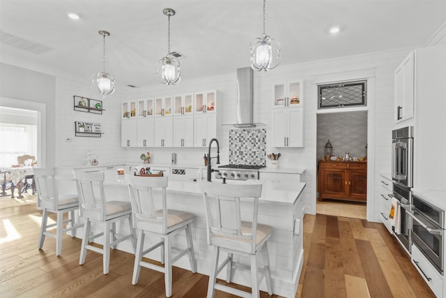 kitchen with light countertops, glass insert cabinets, white cabinetry, wall chimney range hood, and an island with sink