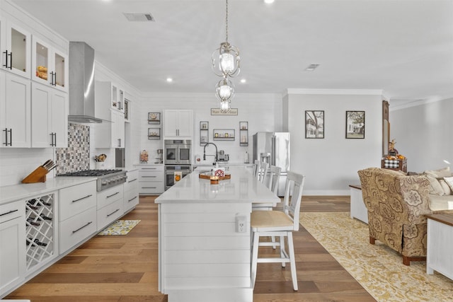 kitchen featuring an island with sink, wall chimney exhaust hood, glass insert cabinets, stainless steel appliances, and light countertops