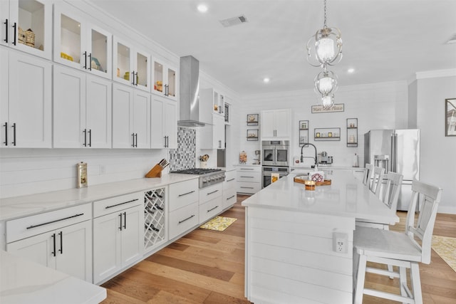 kitchen featuring visible vents, an island with sink, wall chimney exhaust hood, appliances with stainless steel finishes, and glass insert cabinets