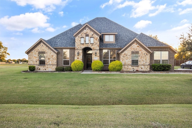 french provincial home with stone siding, brick siding, roof with shingles, and a front yard