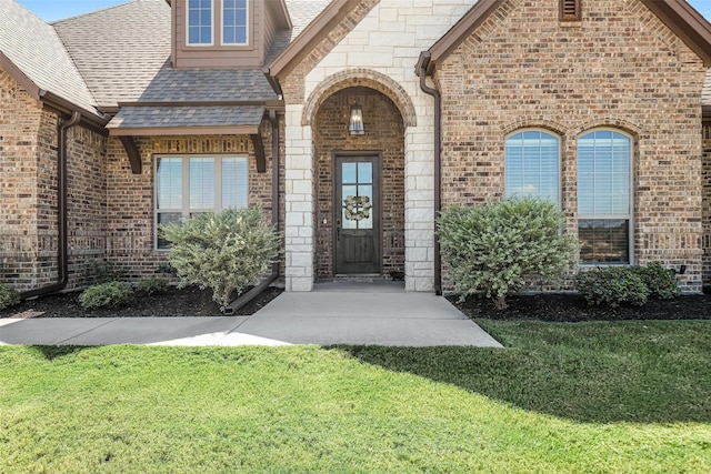 doorway to property with stone siding, brick siding, a lawn, and roof with shingles