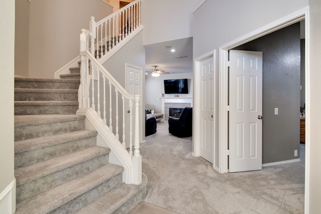 staircase with a towering ceiling, a fireplace, a ceiling fan, and carpet flooring