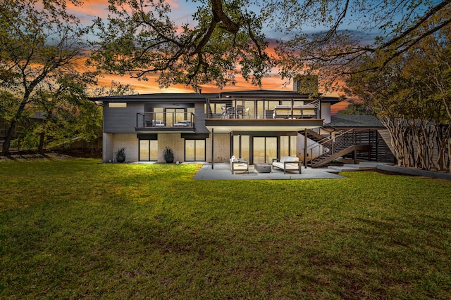 back of property at dusk featuring brick siding, a lawn, a patio area, a balcony, and stairs