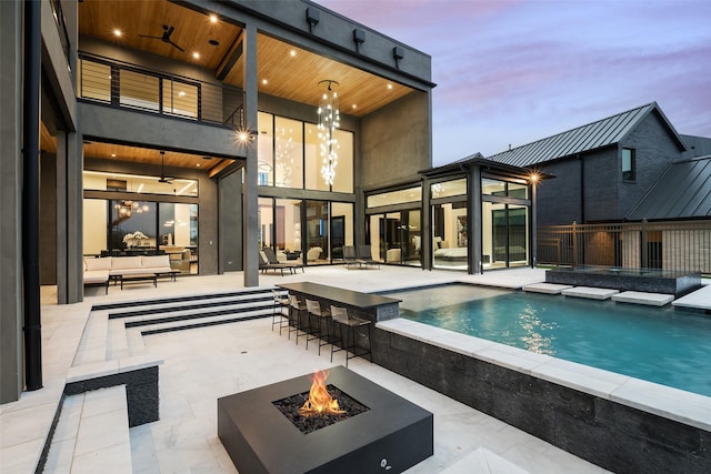 pool at dusk featuring ceiling fan, a patio, a fenced in pool, a bar, and an outdoor living space with a fire pit