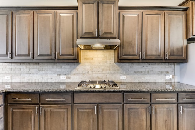 kitchen with stainless steel gas stovetop, decorative backsplash, dark brown cabinets, dark stone counters, and under cabinet range hood