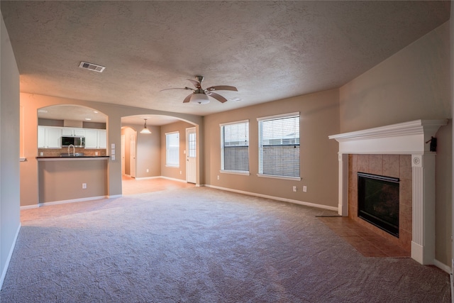 unfurnished living room with baseboards, visible vents, arched walkways, a tiled fireplace, and light colored carpet