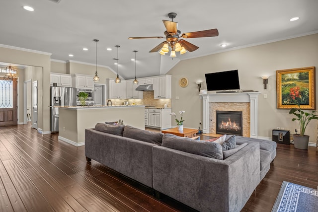 living area with a ceiling fan, ornamental molding, dark wood-type flooring, vaulted ceiling, and a stone fireplace