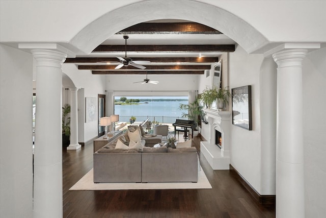 living area featuring a ceiling fan, dark wood-style flooring, a water view, a lit fireplace, and ornate columns