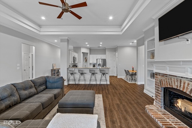 living area featuring built in shelves, dark wood-style flooring, crown molding, a raised ceiling, and a brick fireplace