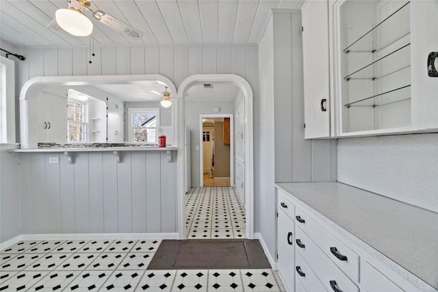 kitchen featuring arched walkways, wood walls, white cabinetry, a ceiling fan, and light countertops