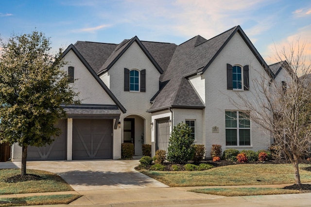 french country inspired facade featuring a garage, brick siding, a shingled roof, driveway, and a front lawn