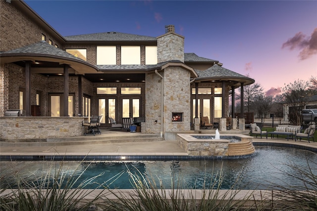 back of house at dusk with brick siding, a patio, a chimney, a pool with connected hot tub, and exterior bar