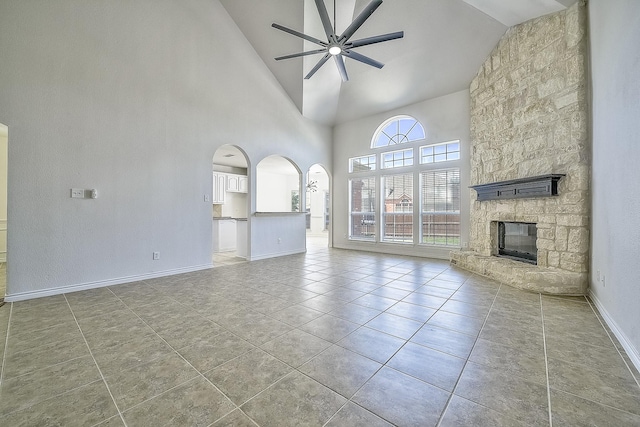 unfurnished living room with arched walkways, light tile patterned floors, a ceiling fan, a stone fireplace, and baseboards