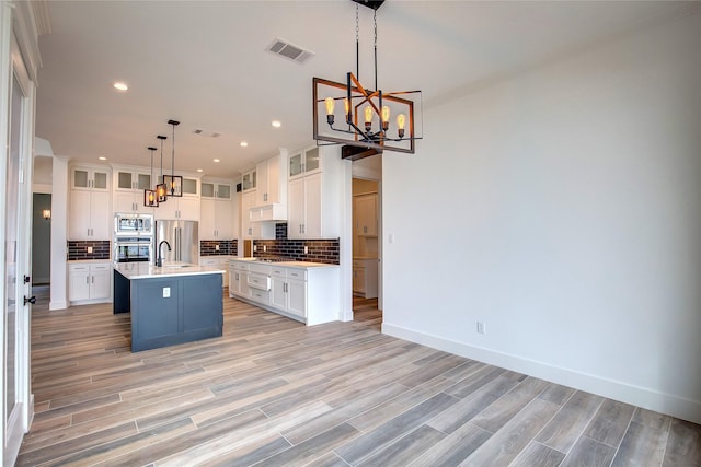 kitchen featuring glass insert cabinets, a kitchen island with sink, stainless steel appliances, light countertops, and pendant lighting