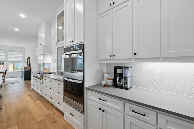 kitchen with stainless steel appliances, backsplash, glass insert cabinets, white cabinetry, and light wood-type flooring