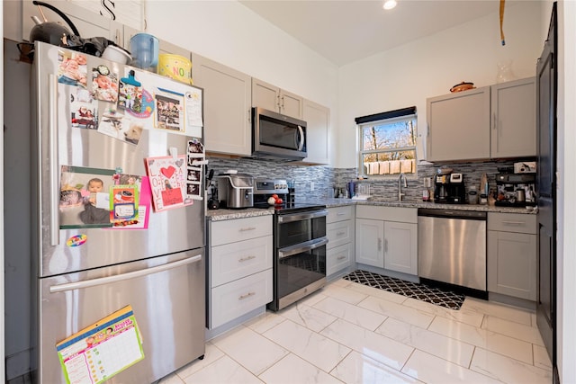 kitchen featuring backsplash, light stone countertops, gray cabinets, stainless steel appliances, and a sink