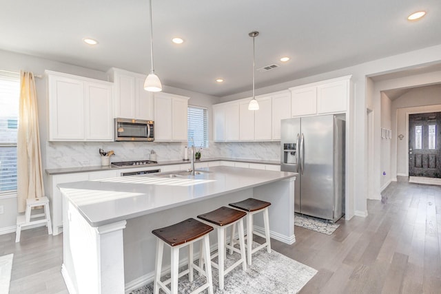 kitchen featuring white cabinets, appliances with stainless steel finishes, decorative light fixtures, a kitchen island with sink, and light countertops