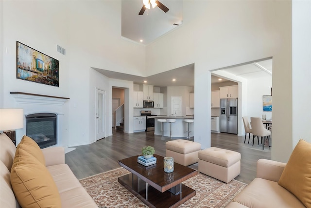 living area featuring visible vents, a glass covered fireplace, ceiling fan, wood finished floors, and a high ceiling