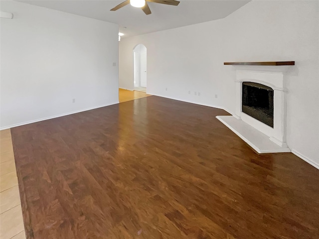 unfurnished living room featuring arched walkways, a fireplace with raised hearth, ceiling fan, wood finished floors, and baseboards