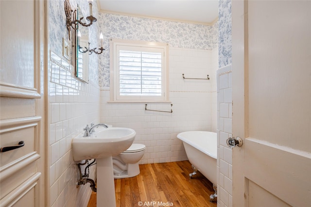 bathroom with wallpapered walls, toilet, a soaking tub, wood finished floors, and tile walls