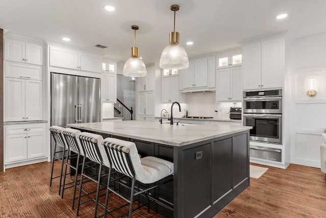 kitchen featuring white cabinetry, appliances with stainless steel finishes, a center island with sink, a warming drawer, and glass insert cabinets