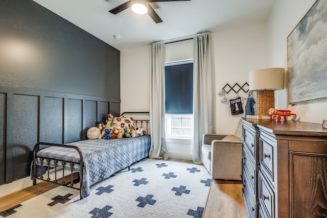 bedroom featuring ceiling fan, wainscoting, light wood-style flooring, and a decorative wall