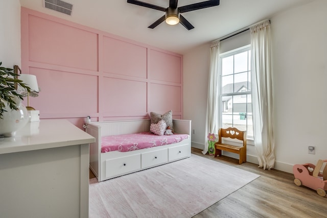 bedroom with ceiling fan, a decorative wall, visible vents, baseboards, and light wood-type flooring