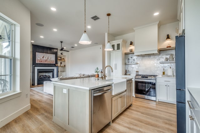 kitchen with stainless steel appliances, light countertops, custom exhaust hood, an island with sink, and glass insert cabinets