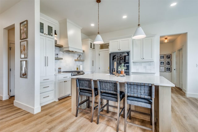 kitchen featuring white cabinetry, light countertops, an island with sink, glass insert cabinets, and custom range hood