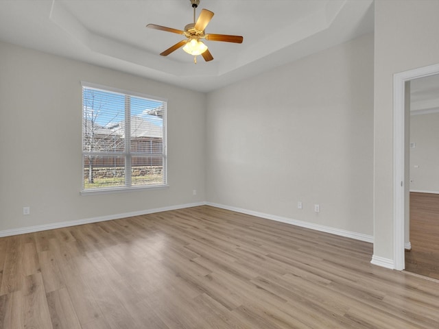 empty room featuring light wood-style flooring, a tray ceiling, ceiling fan, and baseboards